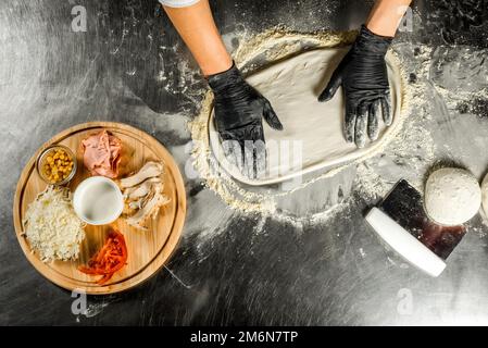 Cooking pizza on a steel table; dough, flour, pizza topping, sauce and chef`s hands in black gloves on the table Stock Photo